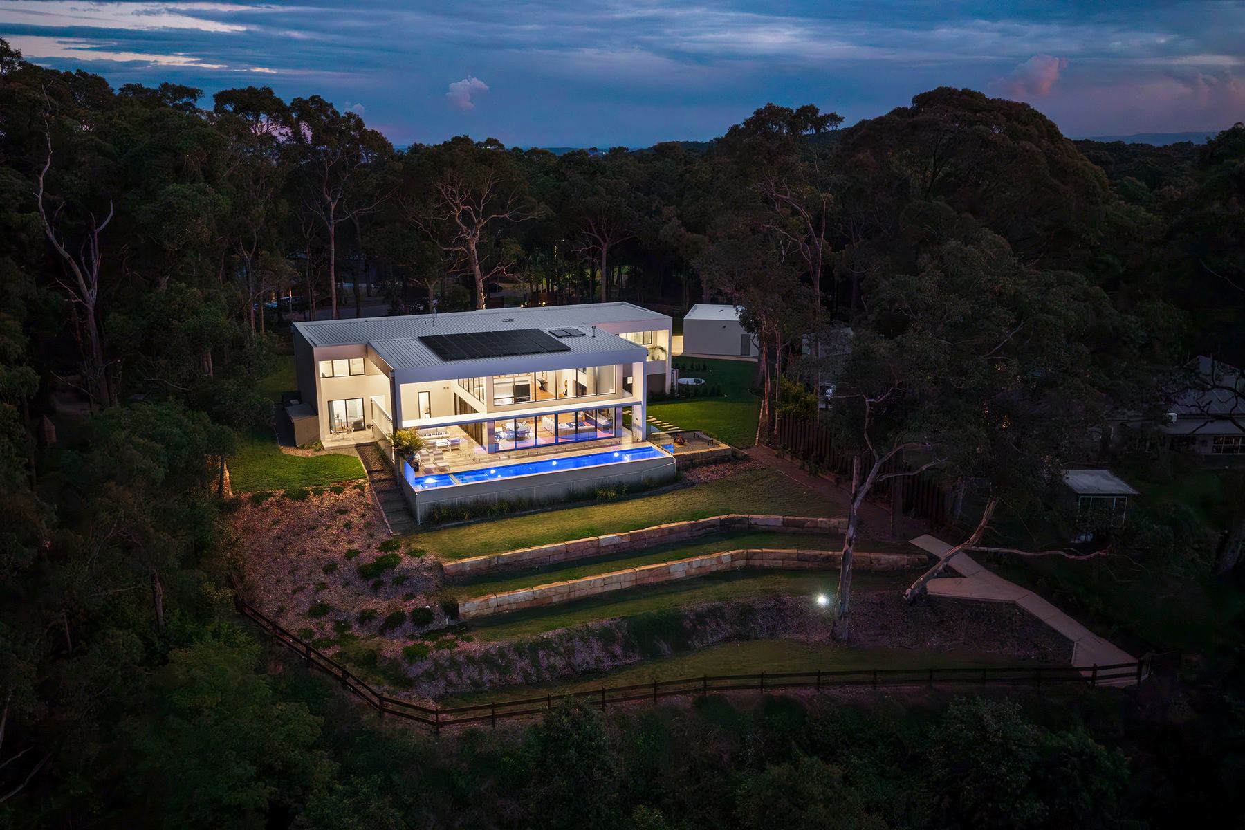 Dusk aerial of a modern acreage residence in Newcastle, warm interior lights and pool glowing against a bushland setting